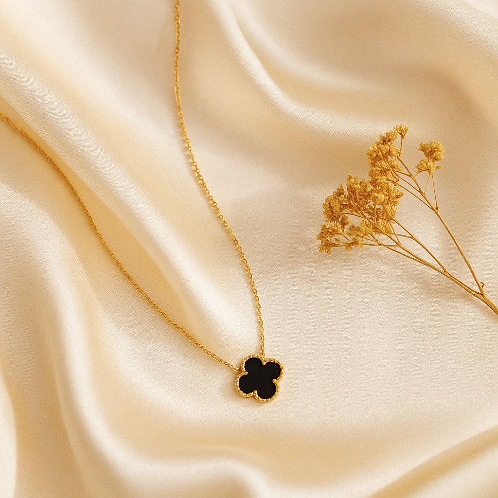 Gold necklace with a black clover pendant on beige fabric with dried flowers.