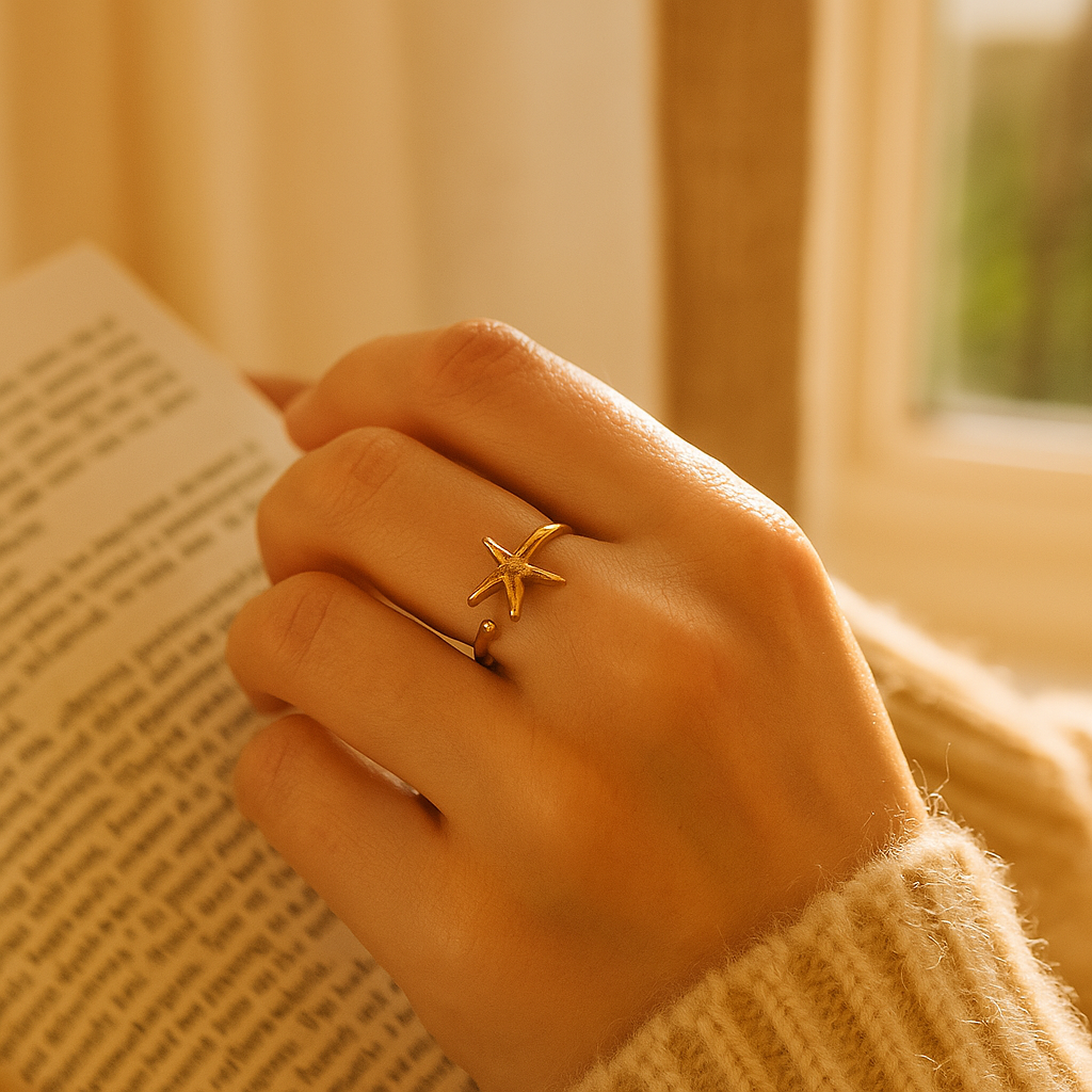 Hand wearing a gold starfish ring holding a book with a warm, blurred background